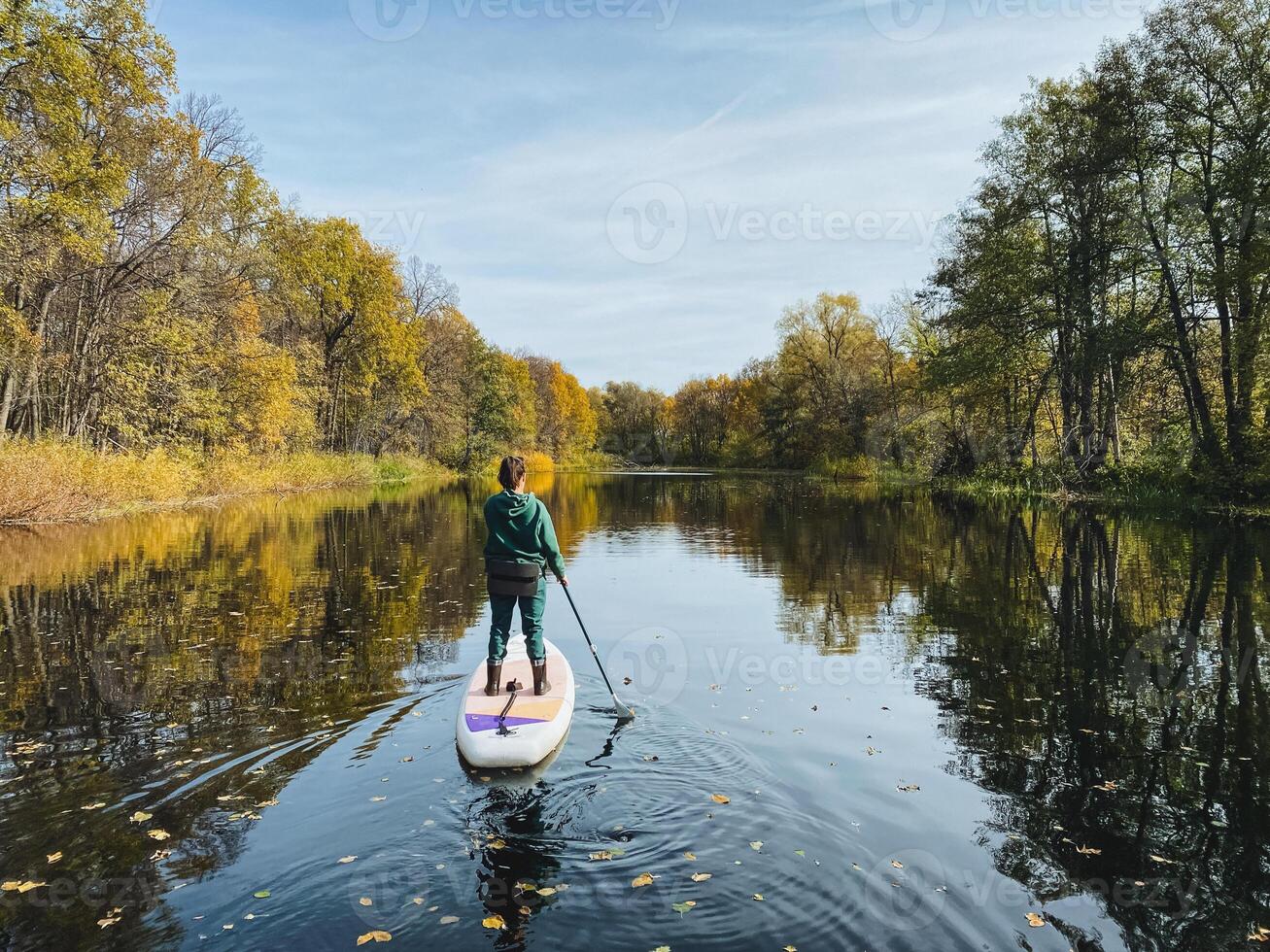 A man is riding a paddle board on a serene river, skillfully navigating the water while taking in the stunning natural scenery surrounding him, from lush trees to the open sky above photo