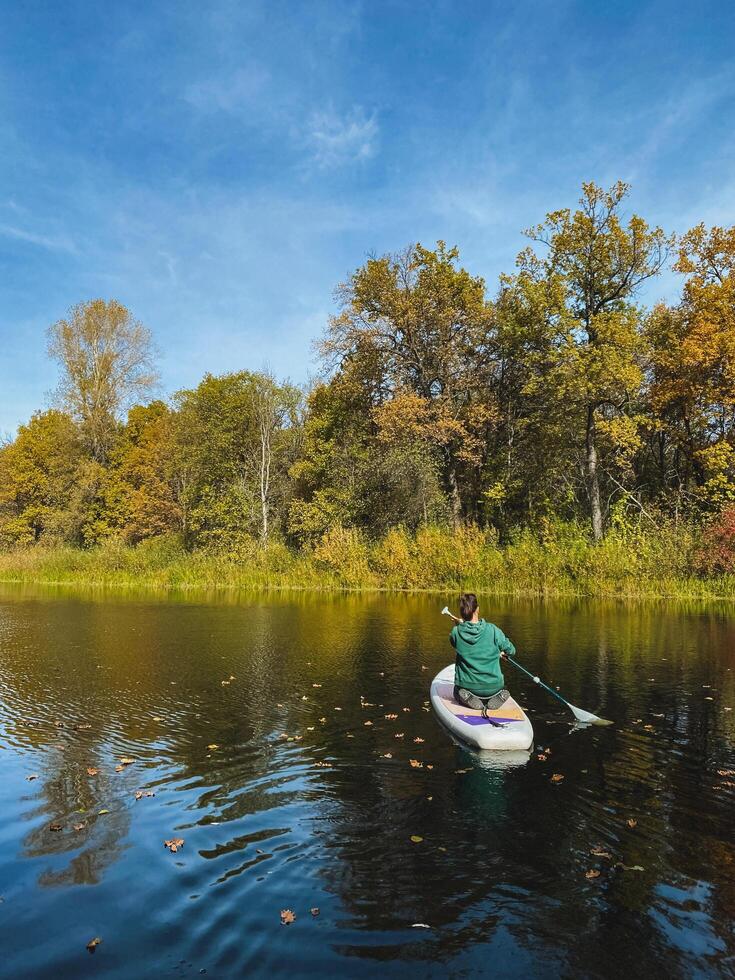 A person is skillfully paddling a paddle board across the serene surface of a beautiful lake, surrounded by lush greenery, under a clear blue sky dotted with soft, fluffy clouds photo