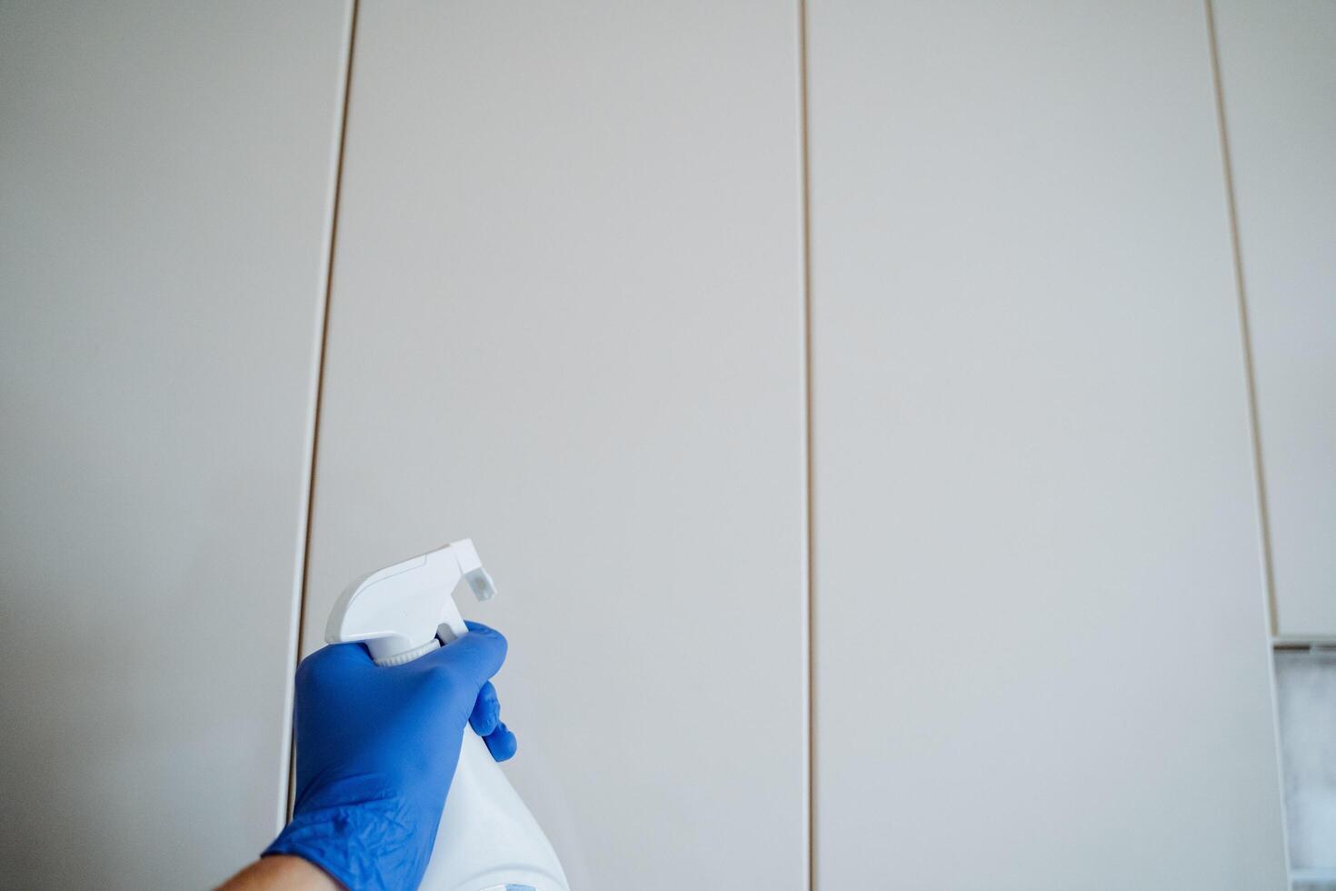 A person wearing vibrant blue gloves confidently holds a spray bottle in front of a pristine white wall, clearly demonstrating their strong commitment to completing a thorough cleaning task photo