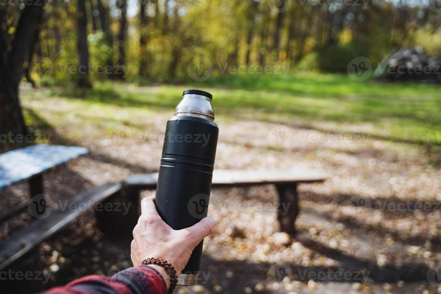 In a picturesque park teeming with vibrant greenery, a person stands proudly holding a thermos bottle in one hand, surrounded by lush trees and soft, inviting grass underfoot photo