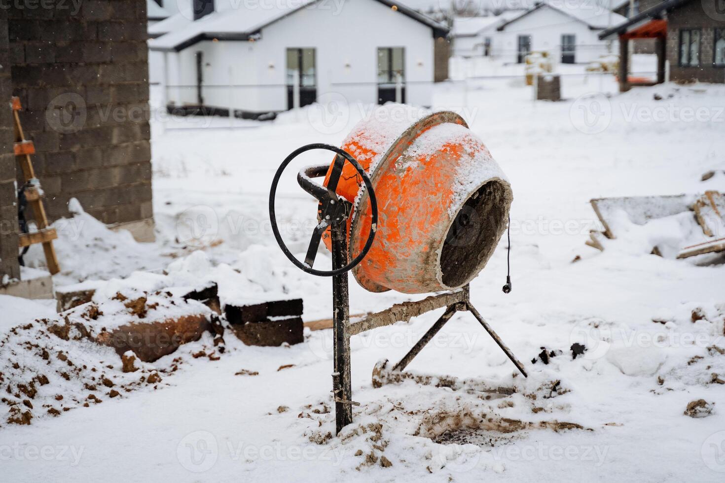 A large concrete mixer is currently sitting idle in the snow on a construction site, surrounded by a white blanket of winter weather conditions, making it appear quite isolated and cold photo