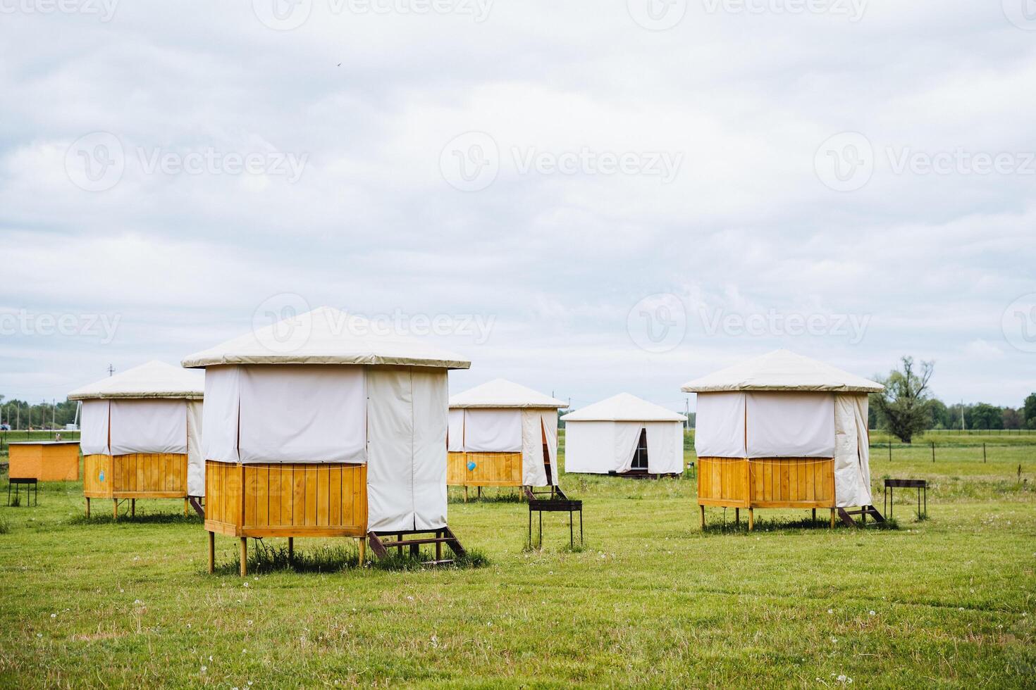 A row of small tents are set up in a grassy field under the open sky with clouds floating by. The natural landscape forms a beautiful backdrop for the event taking place photo