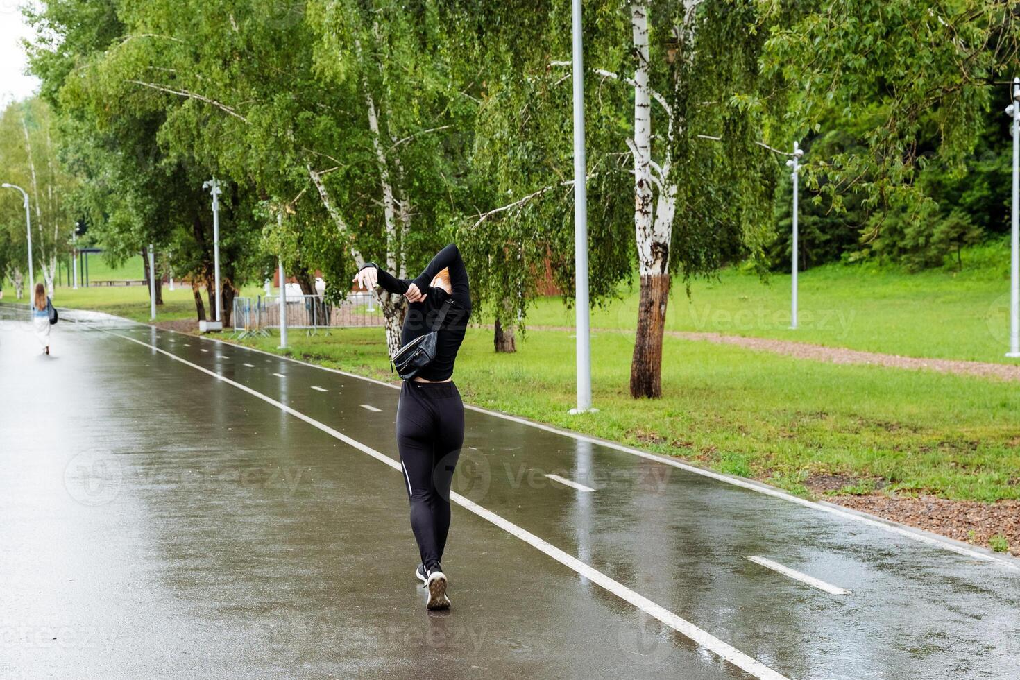 A woman is jogging on a wet road in the rain, surrounded by water, plants, trees, asphalt, grass, and a sidewalk, enjoying her leisure activity and the serene environment photo