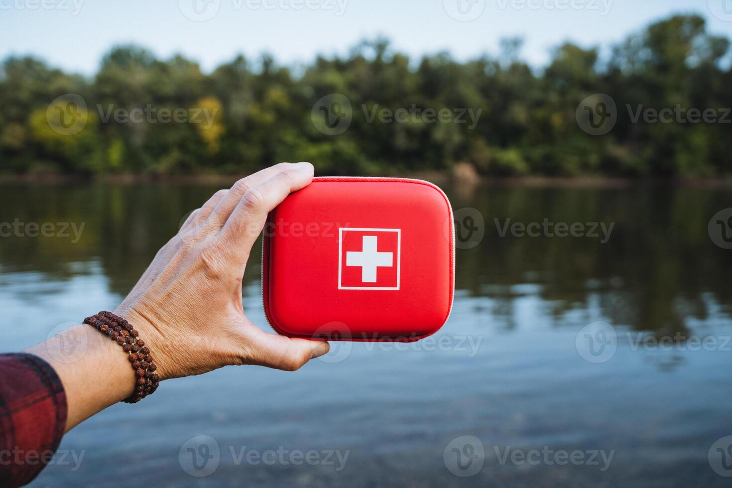 A hand is holding a bright red first aid kit by the water, symbolizing essential emergency preparedness while enjoying the beauty of nature photo
