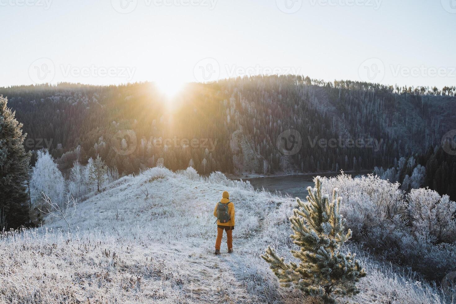 Hiker with backpack walking to meet the sun in the mountains, Sun glare shining at dawn of day, trekking in winter forest, first snow, men's hike alone rear view. photo