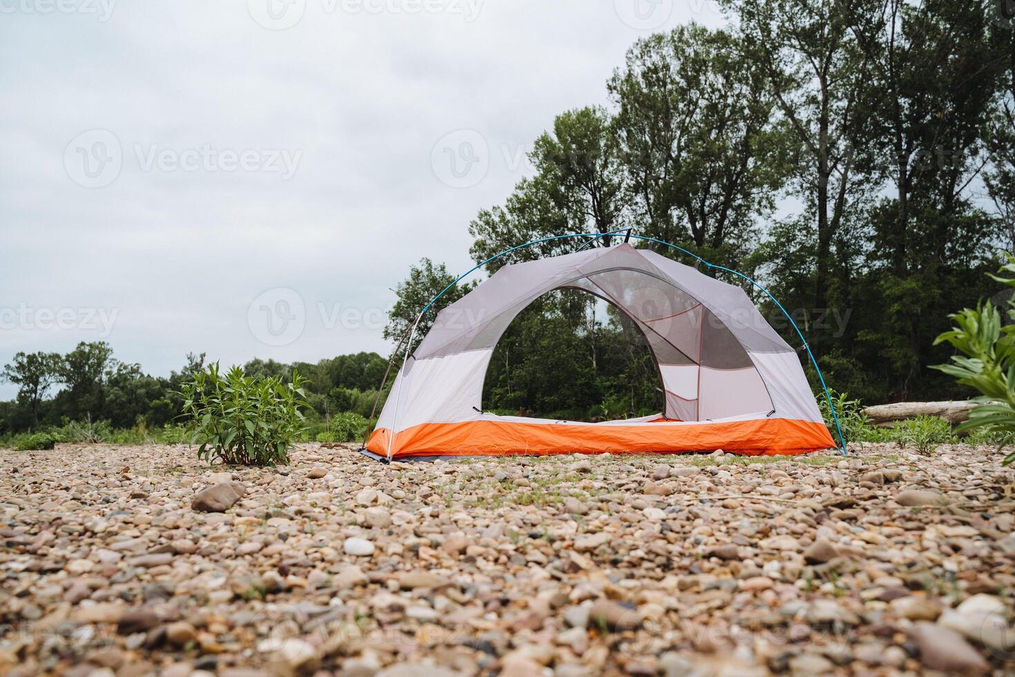 An empty camping tent is set up on a pebble beach with trees in the background on a cloudy day, creating a serene and picturesque landscape for outdoor enthusiasts to enjoy photo