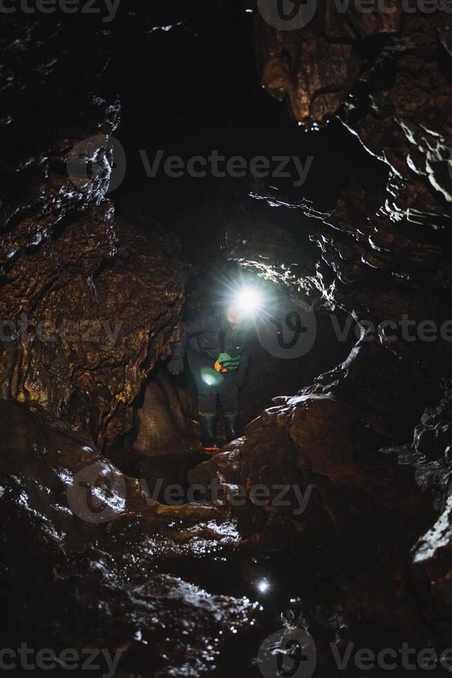 A solitary figure is standing inside a dimly lit cave, wearing a bright light on their head to illuminate the surrounding darkness, surrounded by various natural elements photo