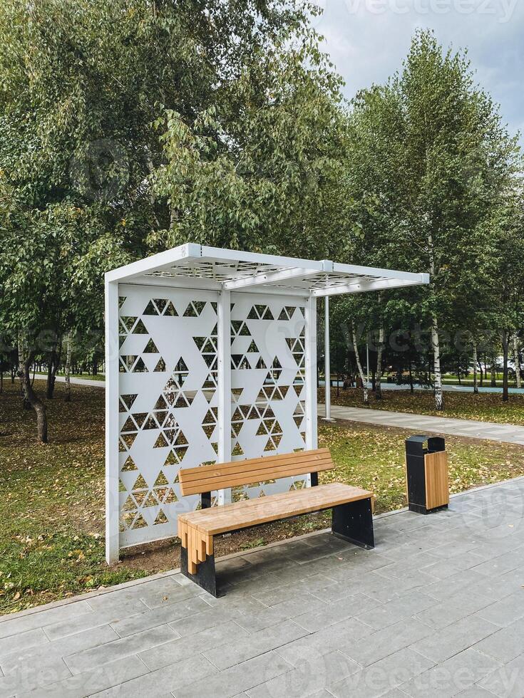 In a vibrant and lively public park, there is a bus stop that features a long comfortable bench along with a conveniently placed trash can for visitors waiting for their transportation photo