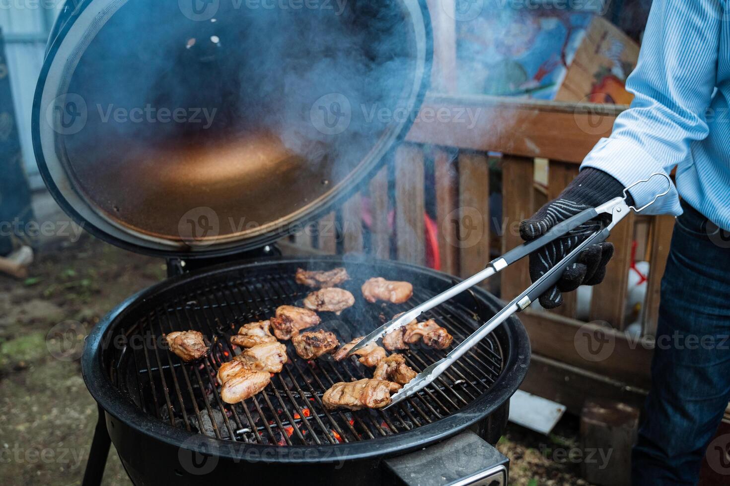 A man grilling chicken on a barbecue grill with tongs, smoke rising in a backyard setting. Keywords grilling, barbecue, outdoor cooking, chicken, smoke, tongs, backyard, summer photo