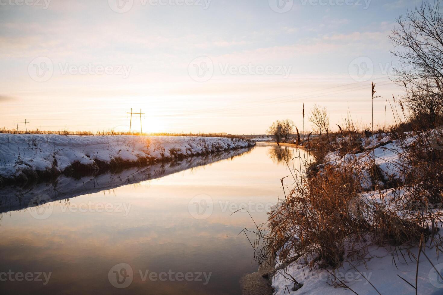 As the day draws to a close, the sun slowly sets over the peaceful, snowy river, casting a soft, warm glow upon the serene winter landscape just moments before nightfall arrives photo