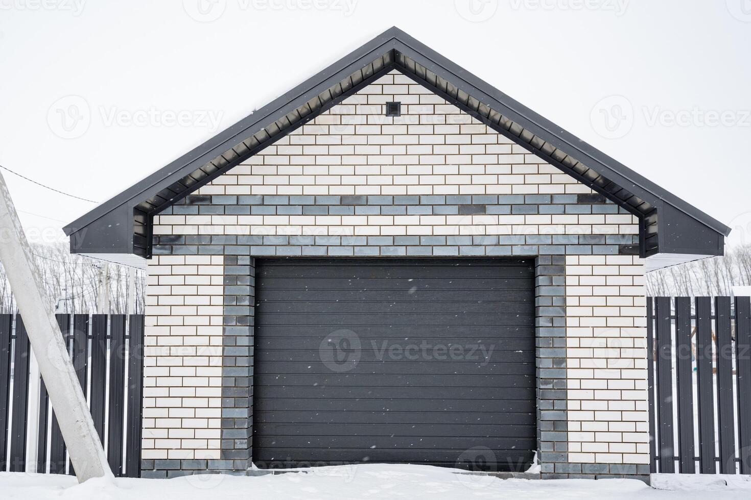A brick garage with a black door and a fence in the snow photo