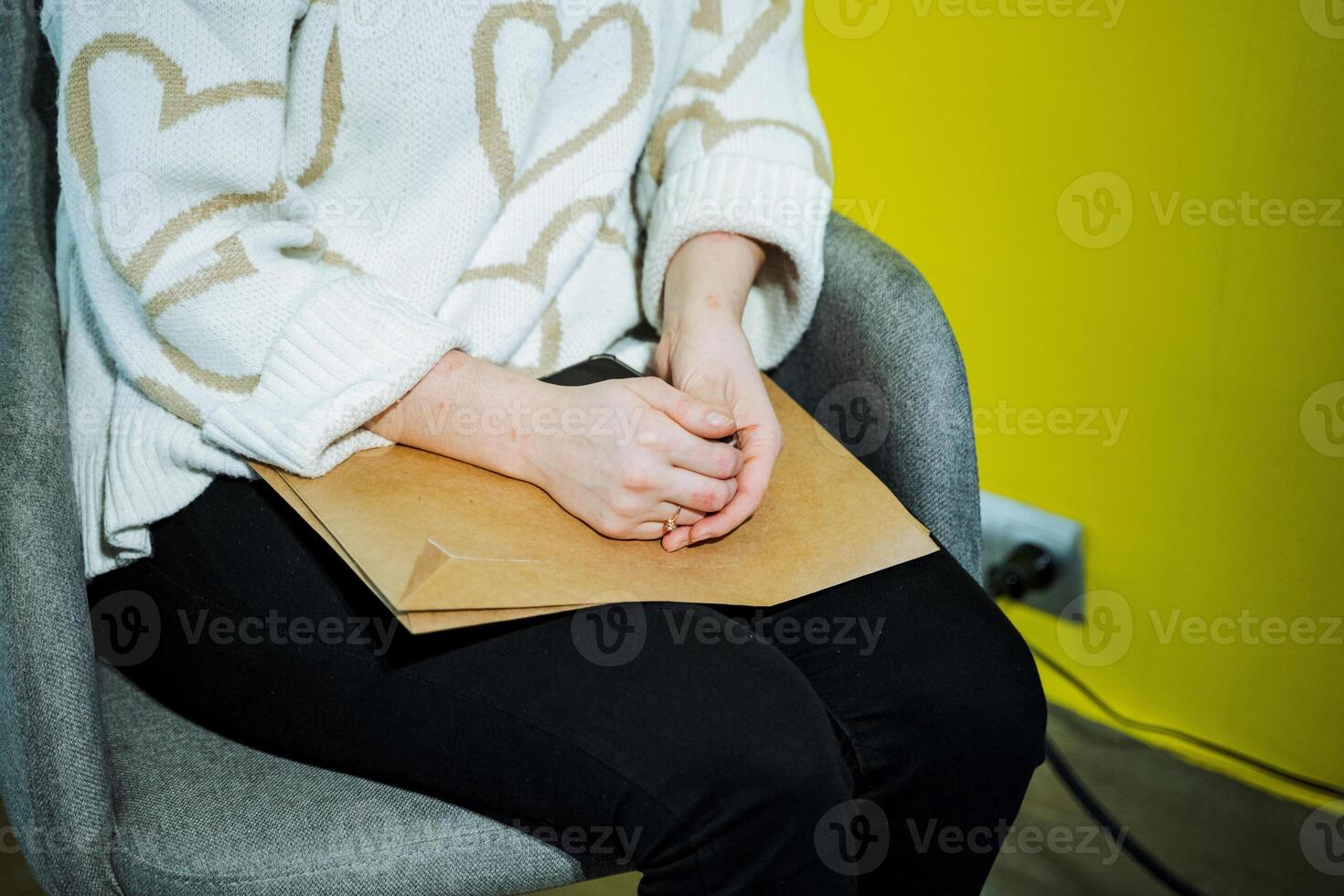 A woman is seated, hands folded, holding a paper, with arms resting on chair photo
