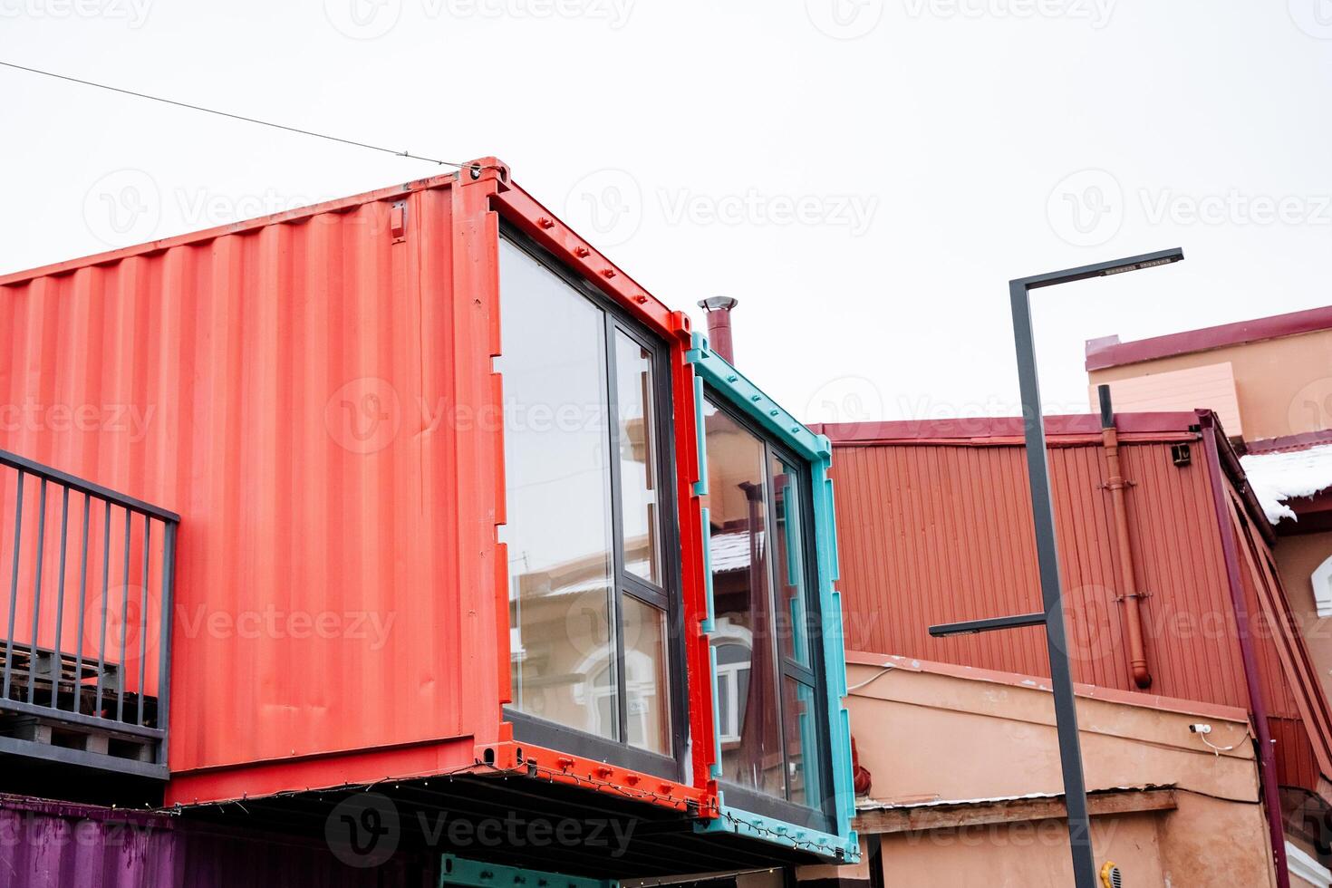 A vibrant red shipping container, which features a balcony, is placed alongside a building, creating an interesting contrast in colors and materials, while enhancing the urban landscape photo