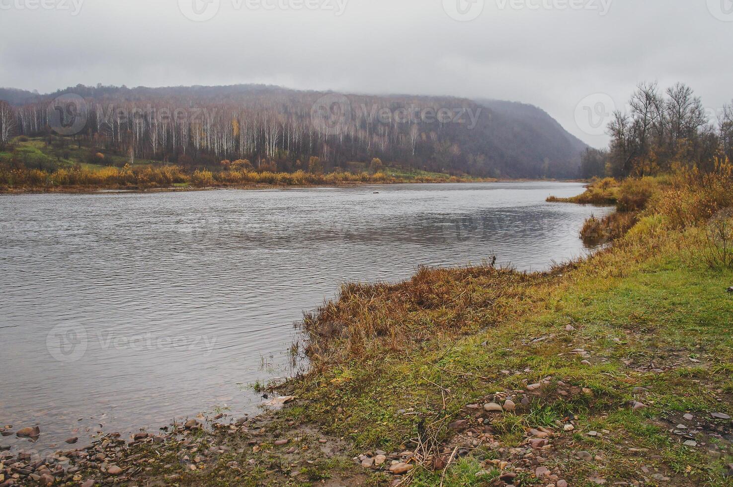 Water flows by a mountain, trees line the shore under a cloudy sky photo