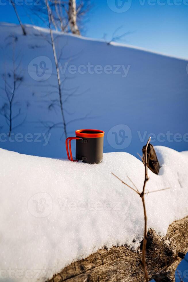 Mug standing on a snow-covered log in the forest, tourist utensils, winter, camp in a snowdrift. photo