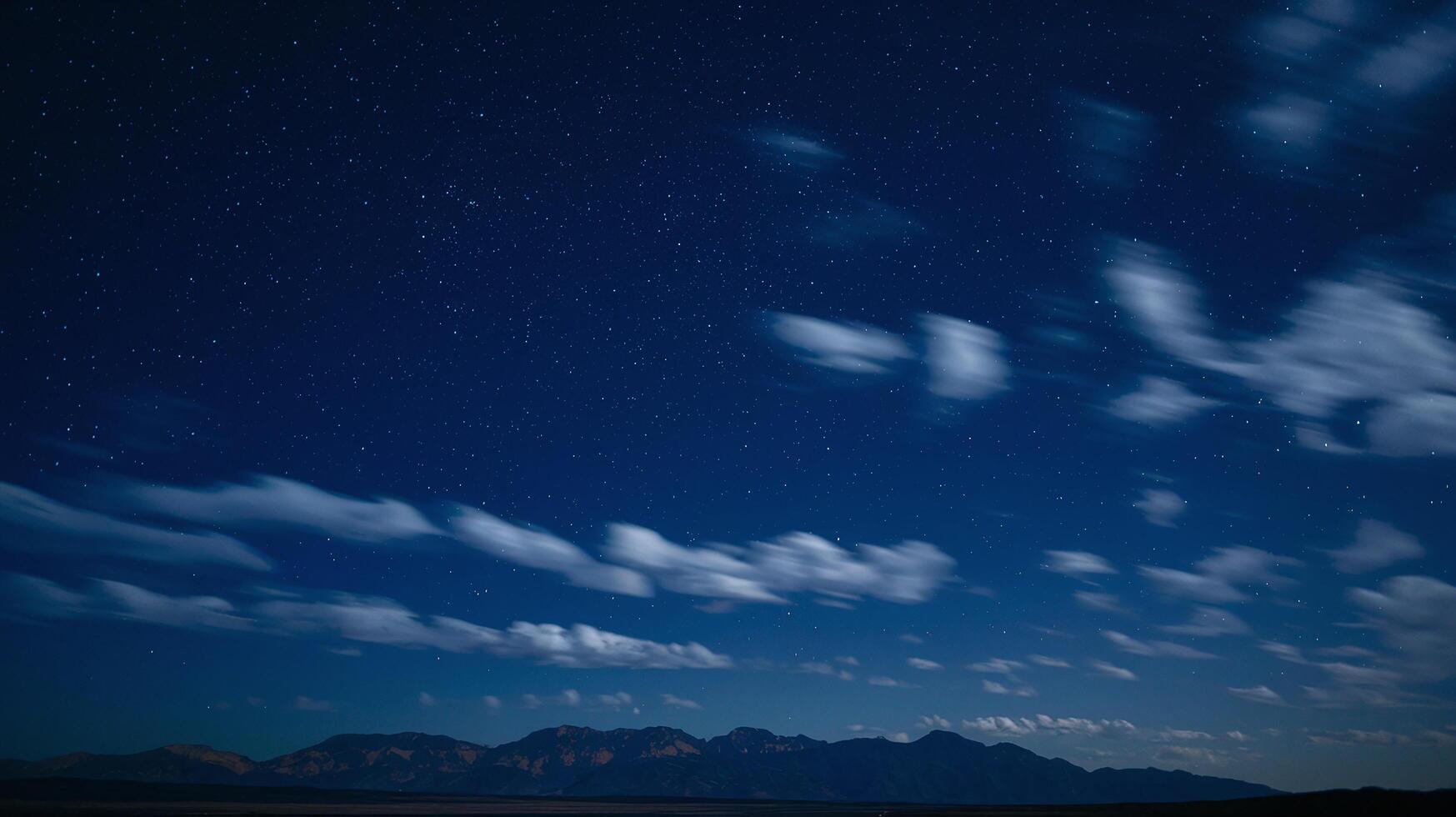 Night Sky with Stars and Moving Clouds Over Distant Mountain Range Under Dark Blue Atmosphere photo