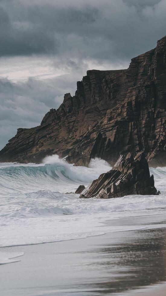 Dramatic Ocean Waves Crashing on Rocky Cliffside with Moody Sky and Sandy Shoreline photo