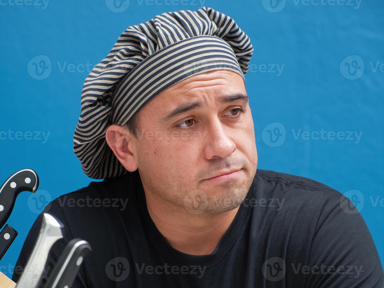 Chef Wearing Striped Hat in Black T-Shirt Posing Thoughtfully with Knives in Blue Background photo
