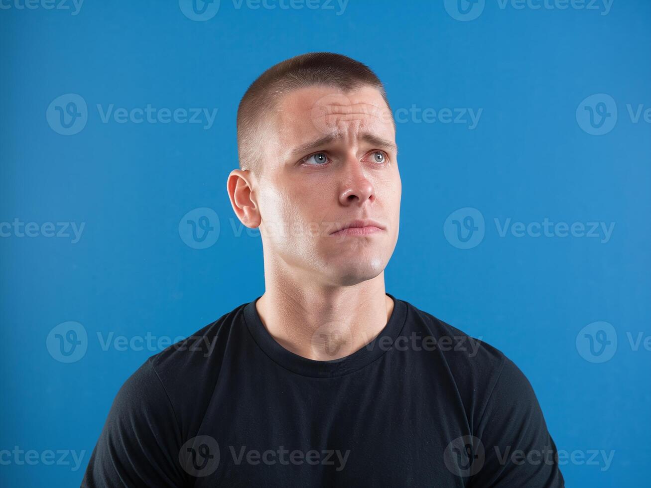 Serious Young Man with Short Hair in Black Shirt Looking Up Against Solid Blue Background photo