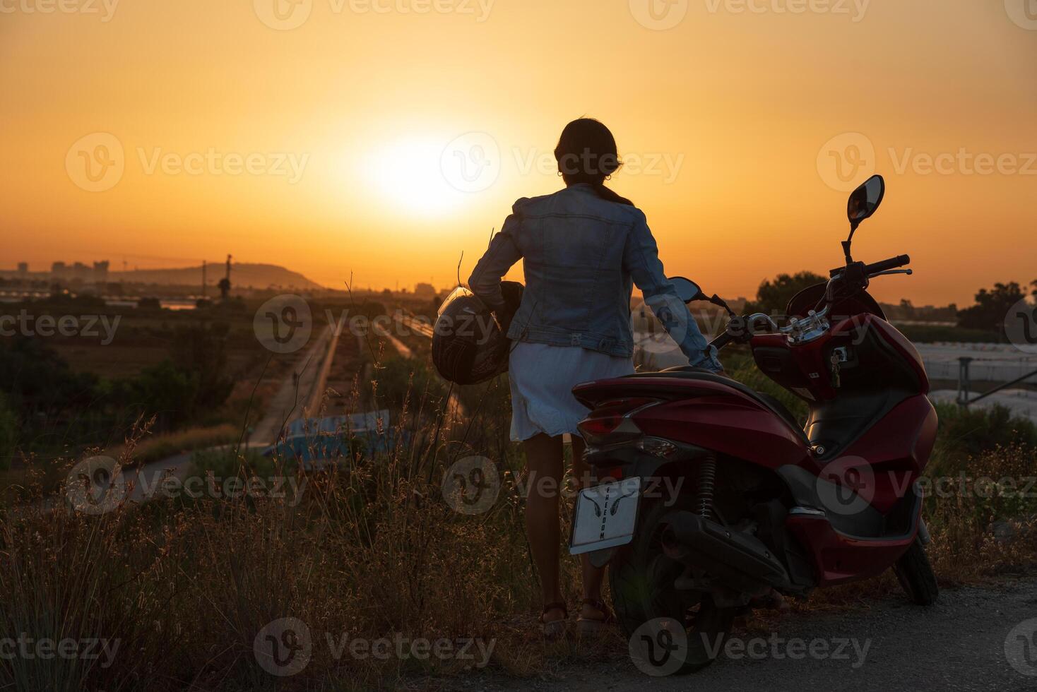 A woman with a helmet in her hand and next to a motorcycle looks out at the path that awaits her on her journey. photo