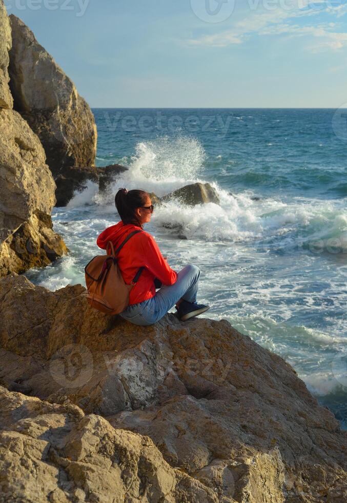 Happy female tourist with a backpack on the top of a mountain, enjoying the sea view, standing on a rock and looking into the distance. The village of Novy Svet in Crimea. Russia photo