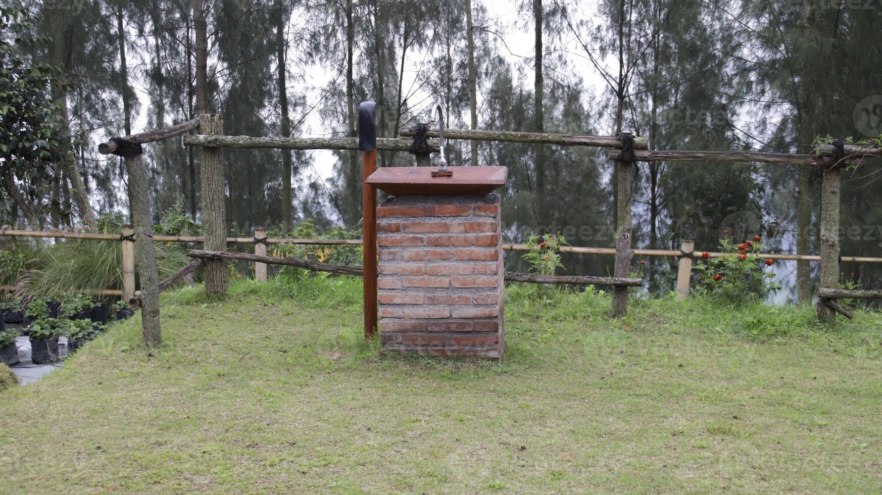 Rustic outdoor brick sink with wooden accents surrounded by lush greenery and natural wooden fences in a scenic rural setting under a cloudy sky photo