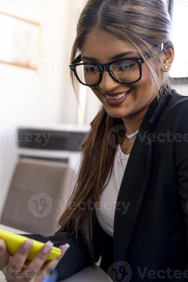 smiling portrait of beautiful business woman with eyeglasses using yellow mobile phone inside modern office photo