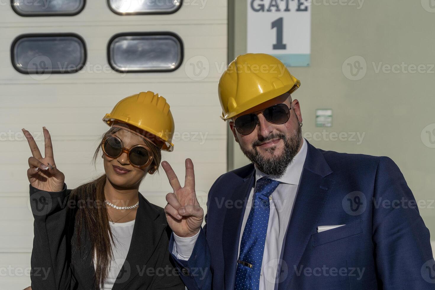 portrait under the sun of two young smiling entrepreneurs, man and woman, showing victory sign wearing yellow safety helmet photo