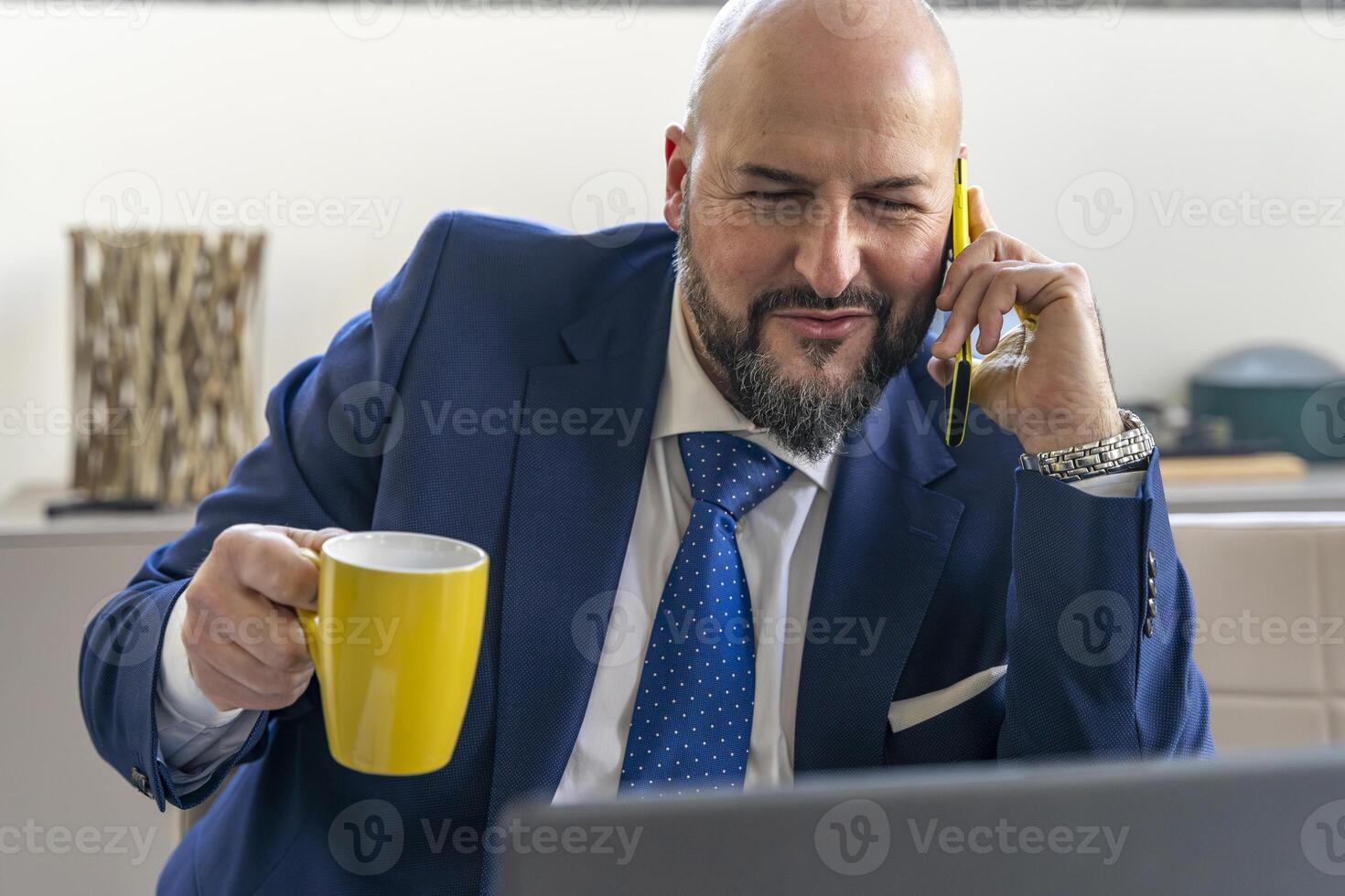 portrait of smiling businessman talking on mobile phone in front of laptop during coffee break in an office photo