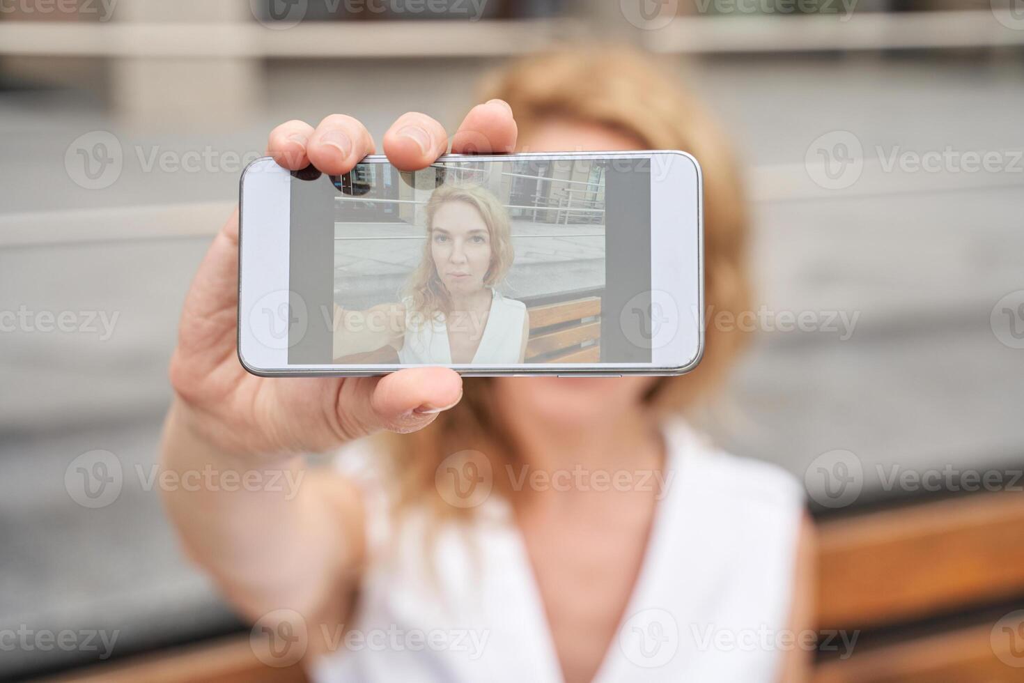 Caucasian female adult taking self-portrait with smartphone, blurred background, cellphone photography focus, woman holding device. photo
