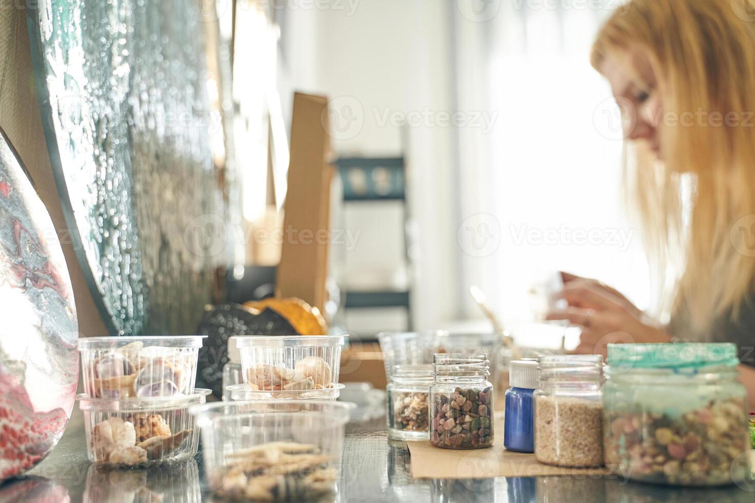 Caucasian female adult creating handmade crafts with assorted stones and containers in sunlit workspace. photo