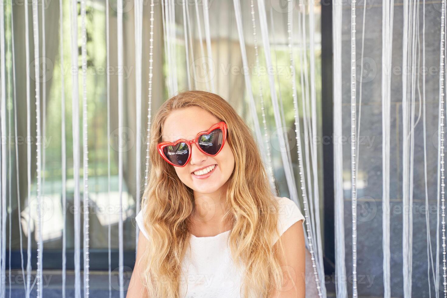 Young caucasian female in heart shaped sunglasses smiling in front of curtain background. photo