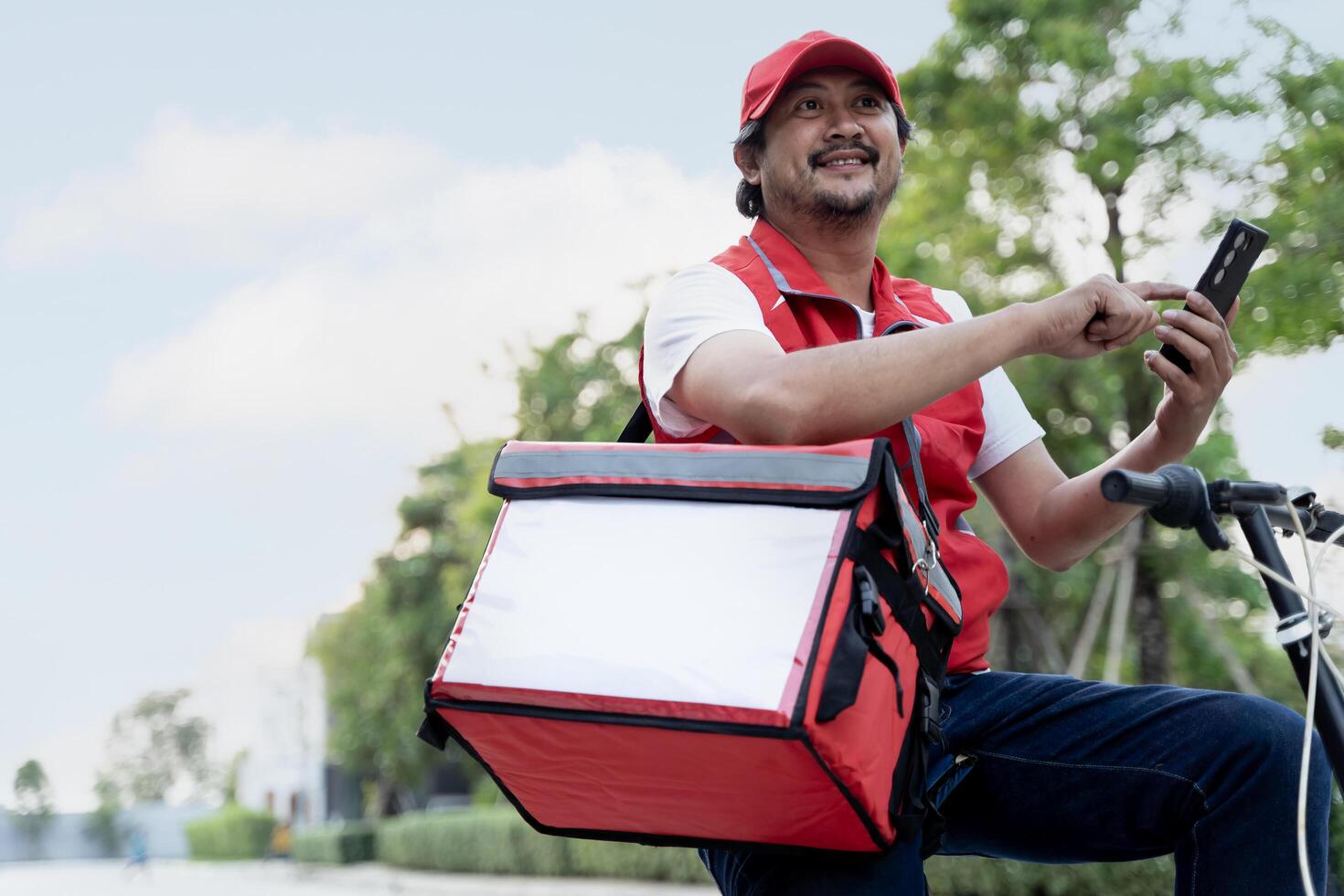 A happy delivery man is sitting on a bicycle and texting on his smartphone. He is carrying a red backpack for delivery on his shoulders and cap. photo