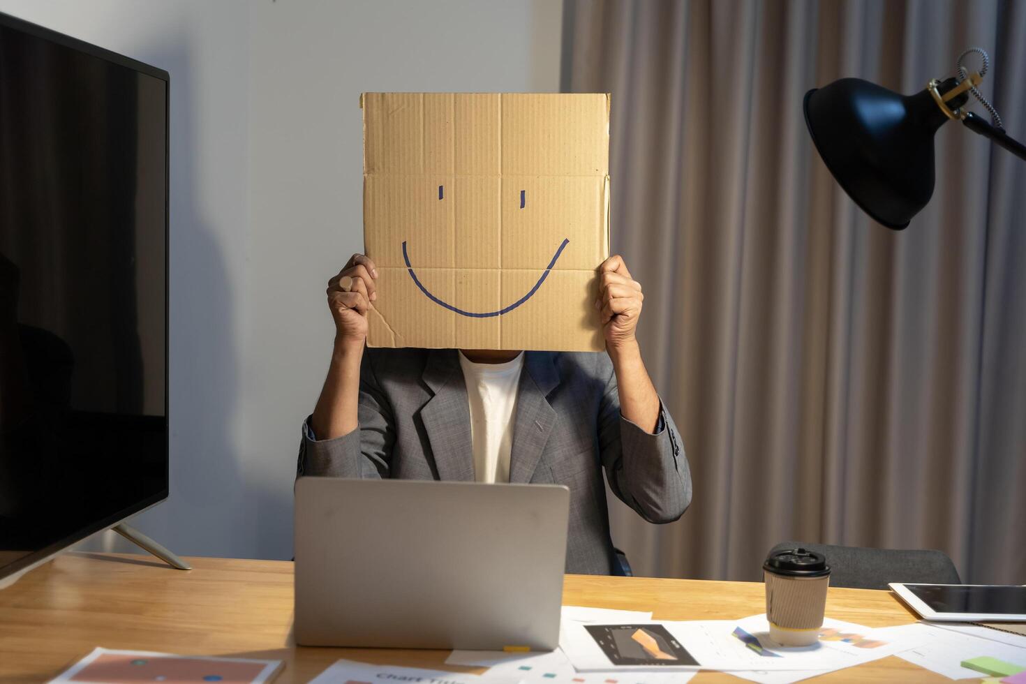 A young businessman is holding a cardboard with a smiley face on it in front of his head, gesturing with a cardboard box. photo
