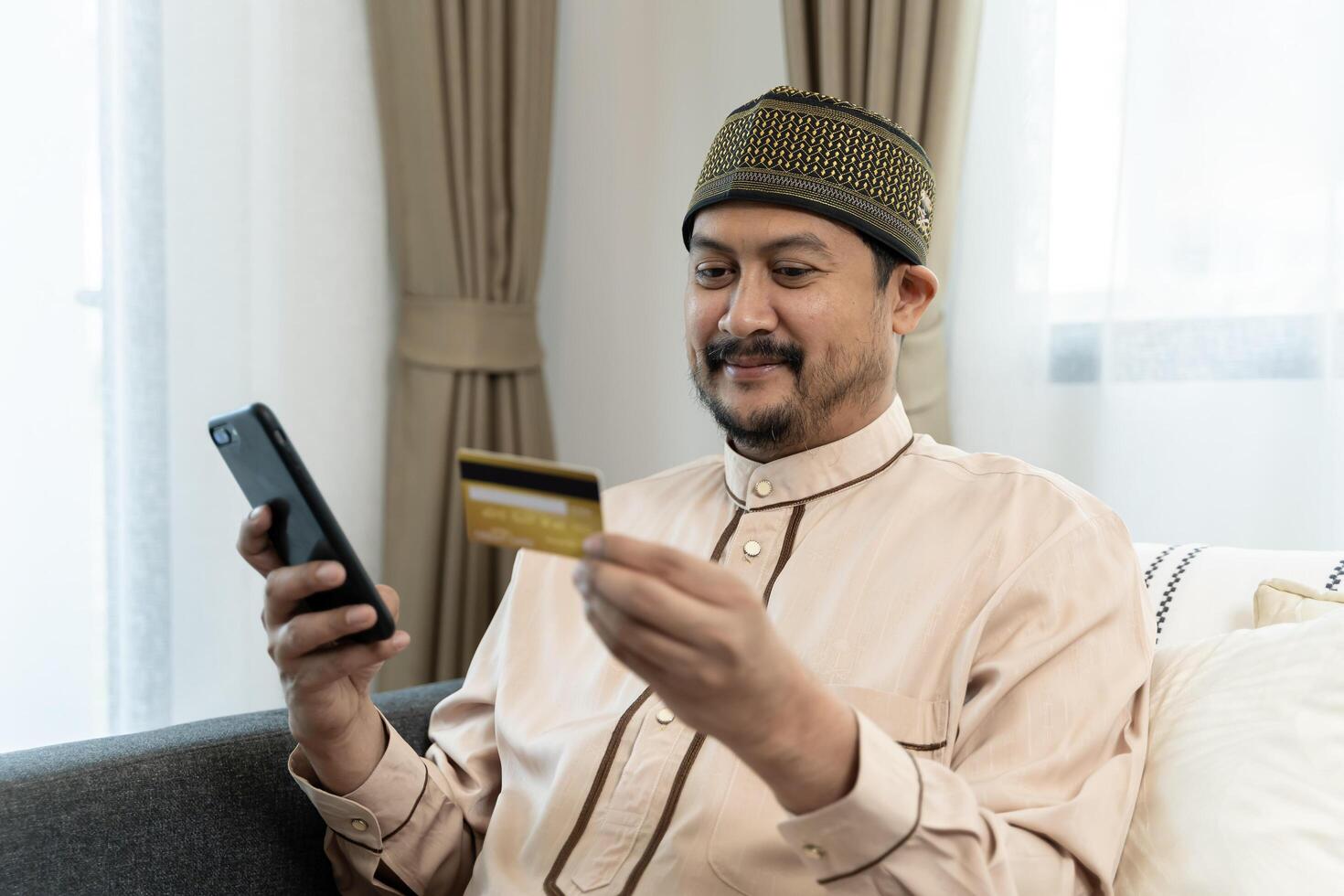 Happy and satisfied man shopping online sitting on sofa at home, hispanic customer holding bank credit card and phone. photo