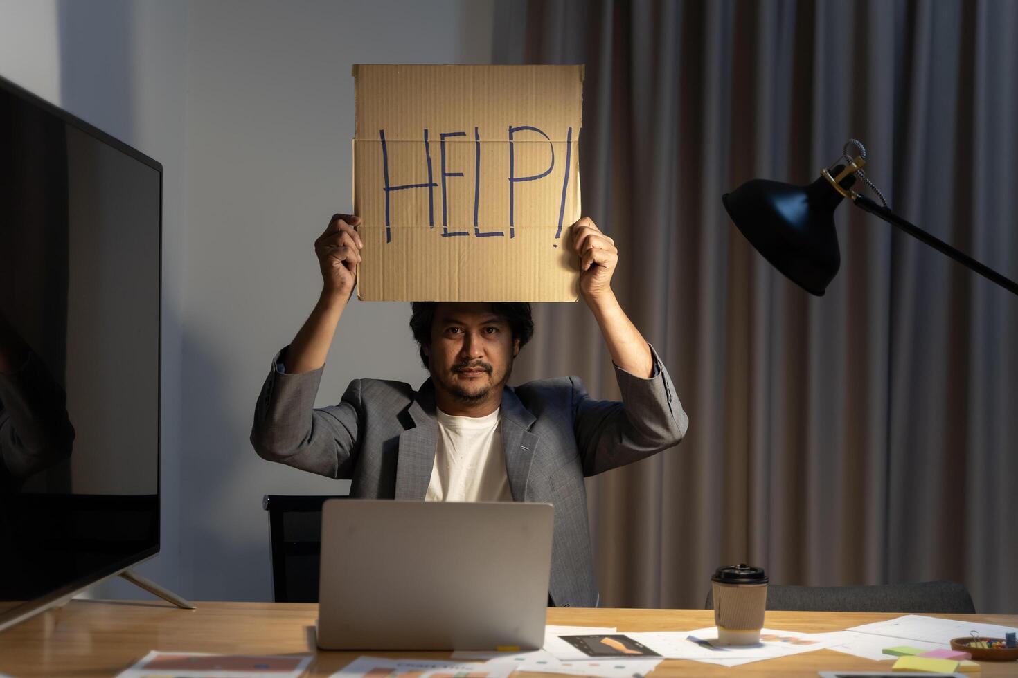 Businessman in suit sitting at office desk working on computer laptop asking for help holding cardboard sign. photo