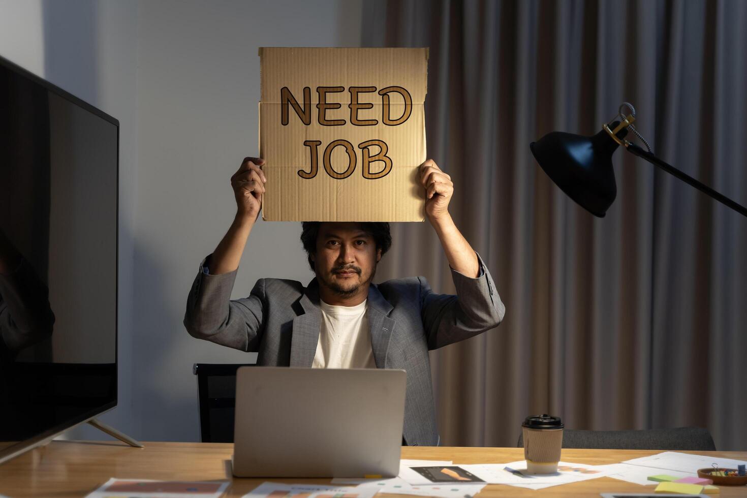 Job hunting. Man sitting at the table in the office, holding a piece of cardboard that says need job in his hands. Hire me concept. photo