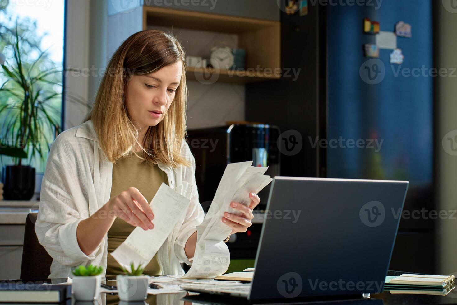 Stressed woman reviewing bills at home. Female organizing receipts, sitting at kitchen table with laptop and paper documents. Concept of budgeting and managing personal finances photo