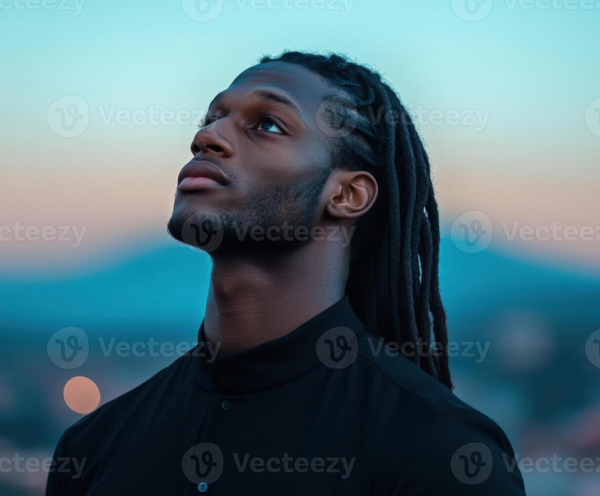 A man with long, dark hair looks up thoughtfully against a soft twilight sky photo