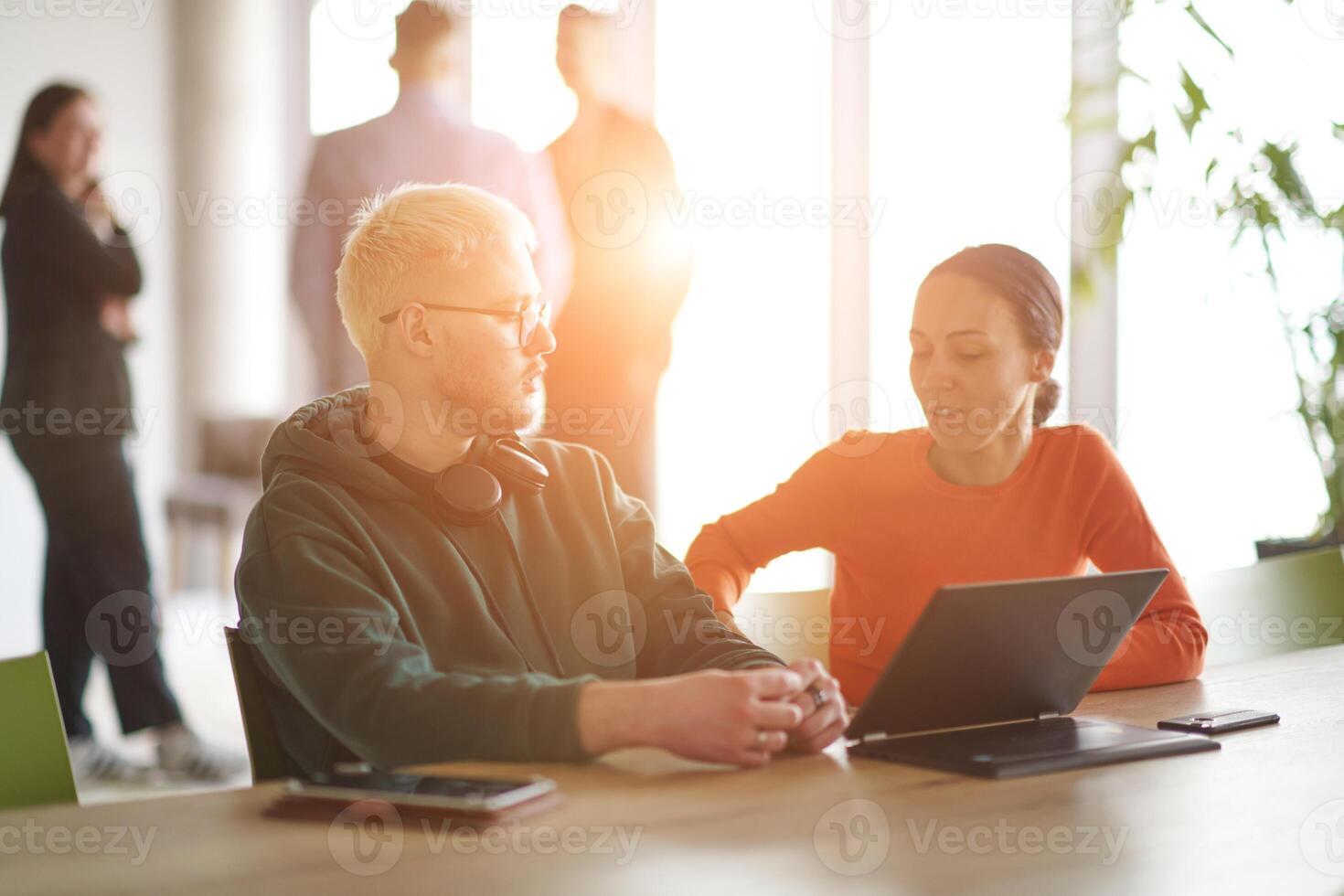 A blond businessman and his colleague sit in an office discussing work while using a laptop to collaborate on tasks photo