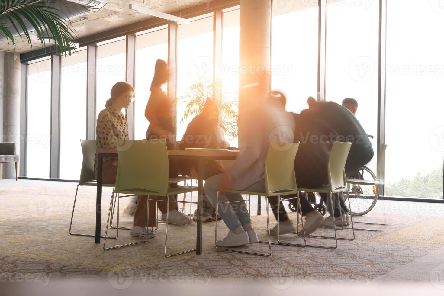 A diverse business team, working urgently around a conference table, collaborates on a meeting while their director, seated in a wheelchair, waits for their input photo