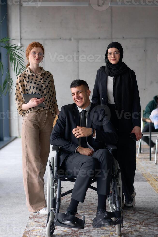 A diverse business team poses confidently, including a smiling director seated in a wheelchair, flanked by two professional female colleagues, one wearing a hijab and the other holding a tablet, in a photo