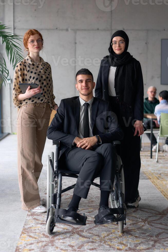 A diverse business team poses confidently, including a smiling director seated in a wheelchair, flanked by two professional female colleagues, one wearing a hijab and the other holding a tablet, in a photo