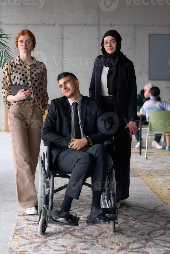 A diverse business team poses confidently, including a smiling director seated in a wheelchair, flanked by two professional female colleagues, one wearing a hijab and the other holding a tablet, in a photo