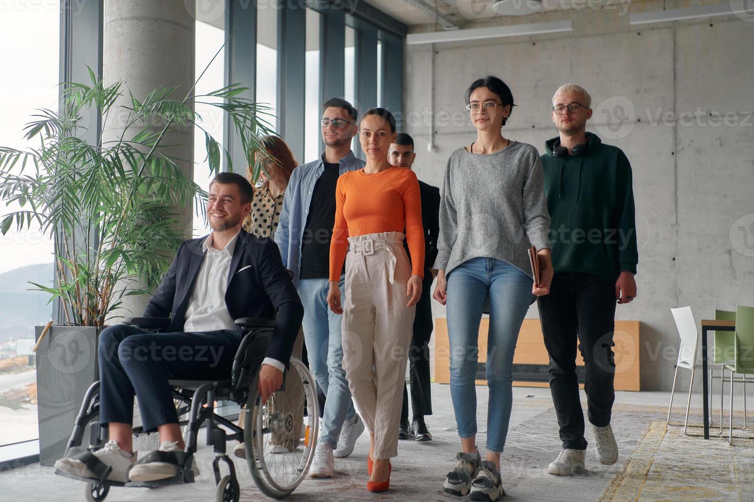 A team ofbusiness professionals moves through a spacious, modern office, assisting their leader, a businessman in a wheelchair, as they collaborate and navigate the workplace together. photo