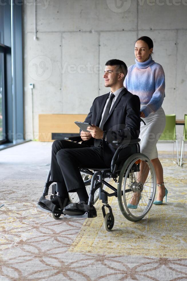 In a professional office setting, a businesswoman helping her director, who is sitting in a wheelchair and using a tablet, while their colleagues collaborate in the background. photo