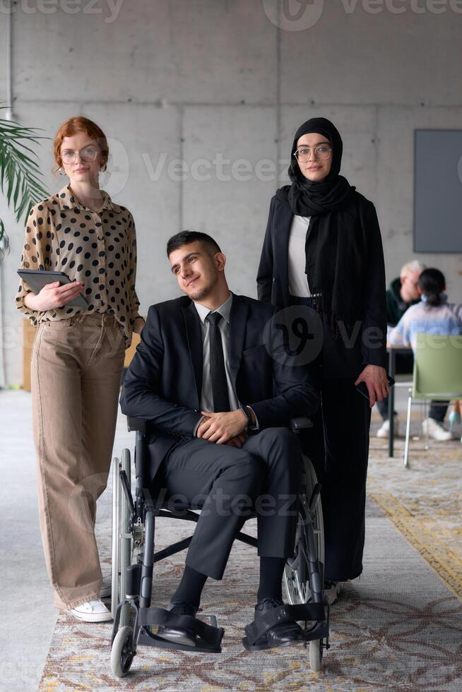 A diverse business team poses confidently, including a smiling director seated in a wheelchair, flanked by two professional female colleagues, one wearing a hijab and the other holding a tablet, in a photo