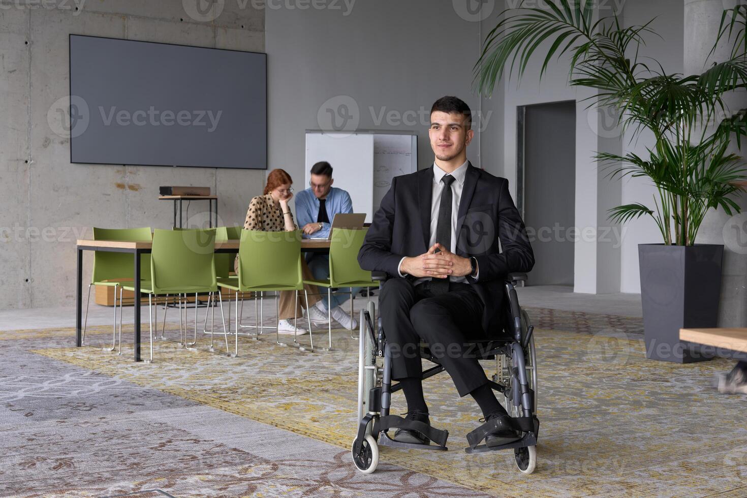 Business Director in a Suit Using a Wheelchair with Colleagues Conducting a Meeting Behind Him photo