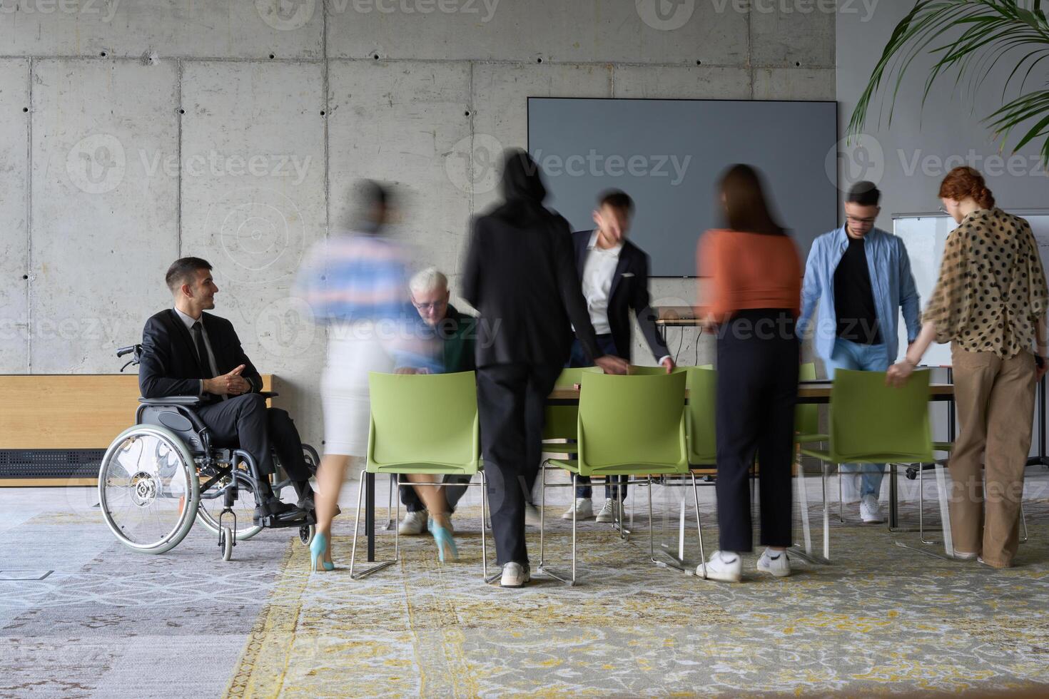 A diverse business team, working urgently around a conference table, collaborates on a meeting while their director, seated in a wheelchair, waits for their input photo