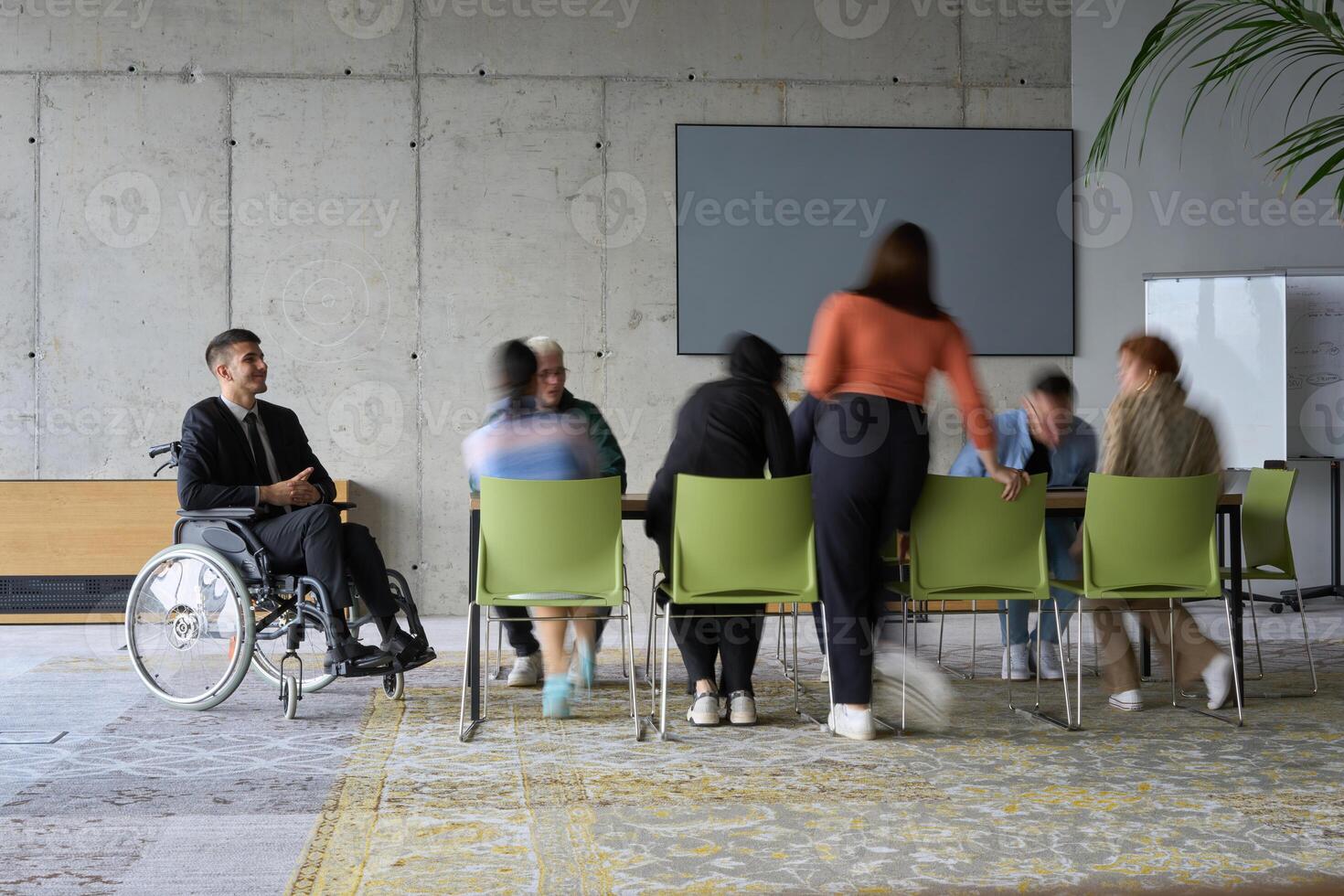 A diverse business team, working urgently around a conference table, collaborates on a meeting while their director, seated in a wheelchair, waits for their input photo